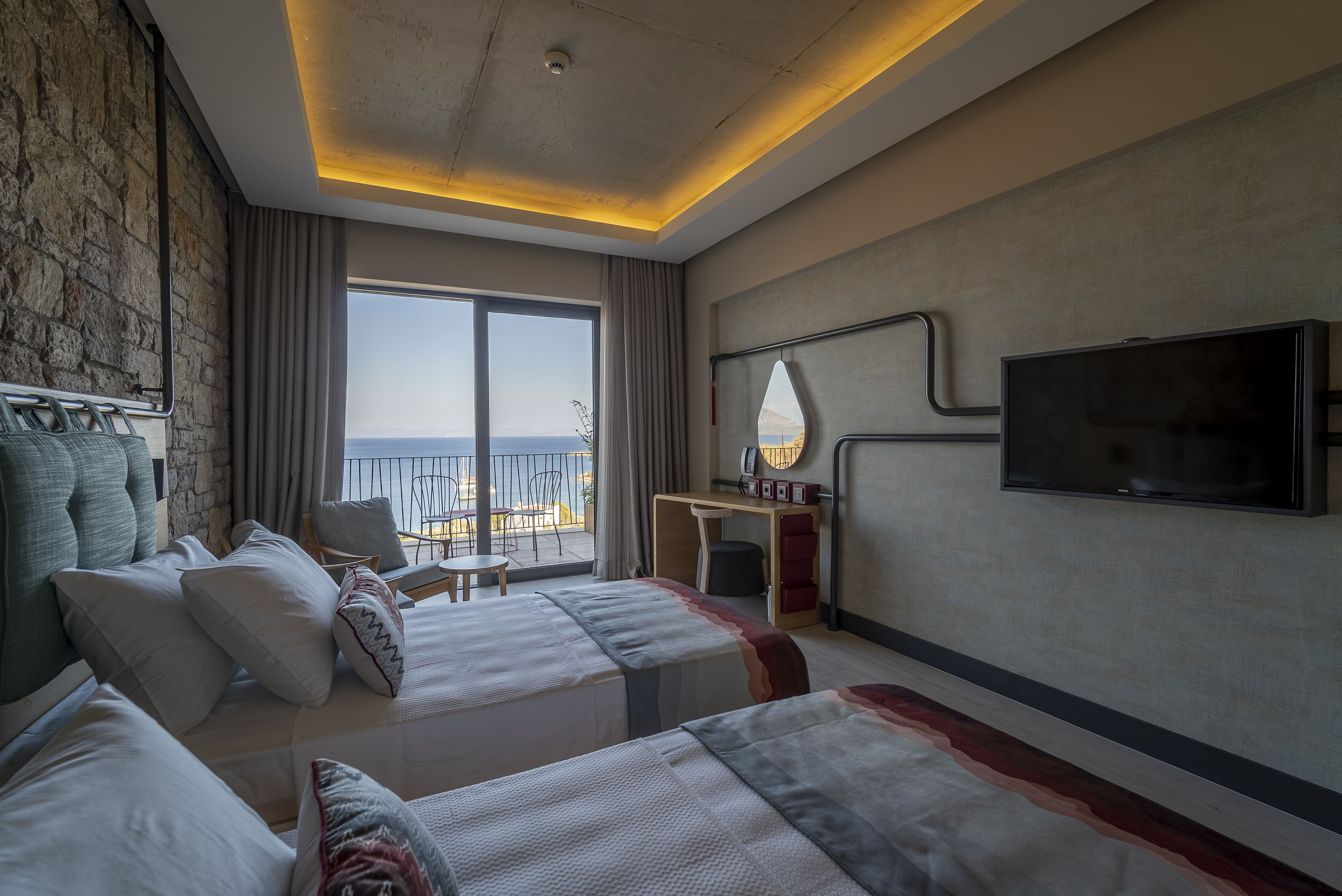 A hotel room with a large bed, a desk, a chair, and a TV. There is a balcony with a view of the sea.