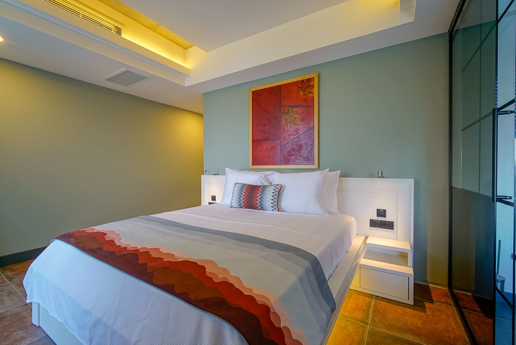 Habitación de hotel con una cama grande, una mesita de noche, una obra de arte colgada en la pared y un panel de vidrio.