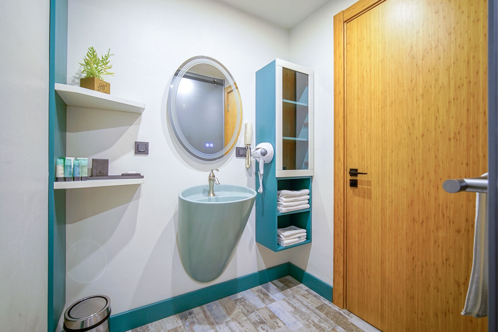 A bathroom with a round mirror, a sink, a hairdryer, and shelves with toiletries.
