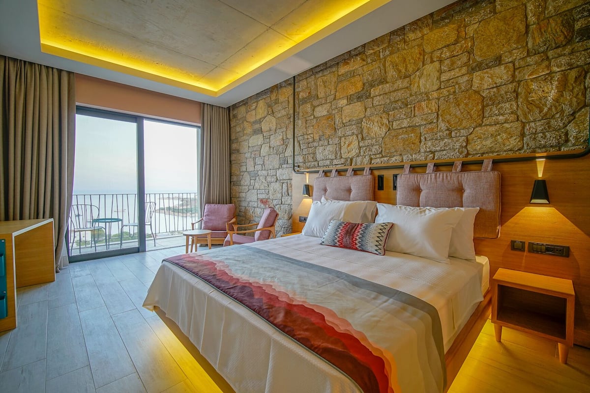 Habitación de hotel con una cama grande, pared de piedra, balcón con sillas y vista al mar.