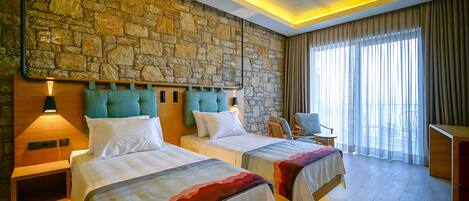 Une chambre d’hôtel avec deux lits, un mur en pierre, une tête de lit en bois, une table de chevet, une lampe, une chaise, une petite table et une grande fenêtre avec des rideaux.