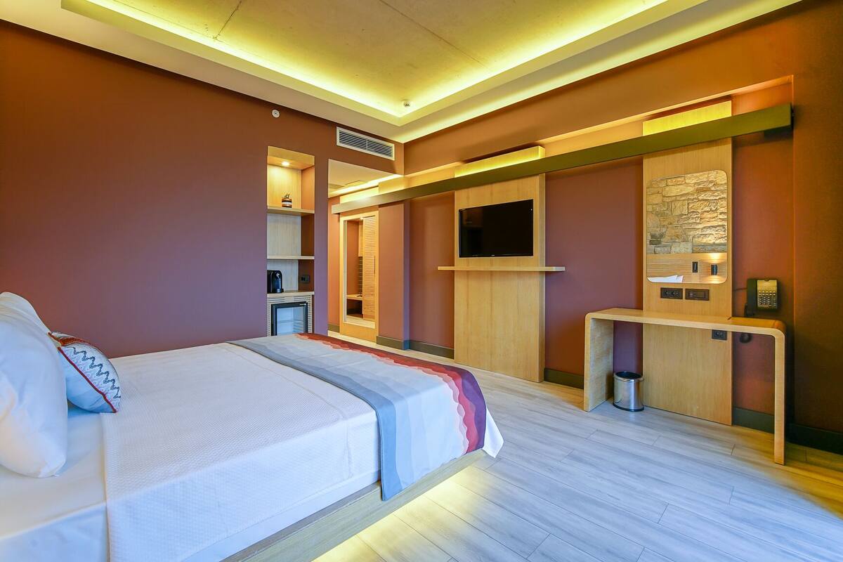 Habitación de hotel moderna con cama, escritorio, televisión y una kitchenette pequeña.