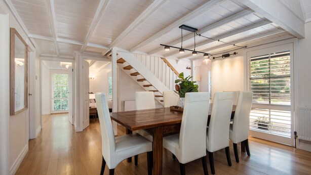 Comedor con mesa de madera y sillas blancas, una escalera y vista a la vegetación por la ventana.