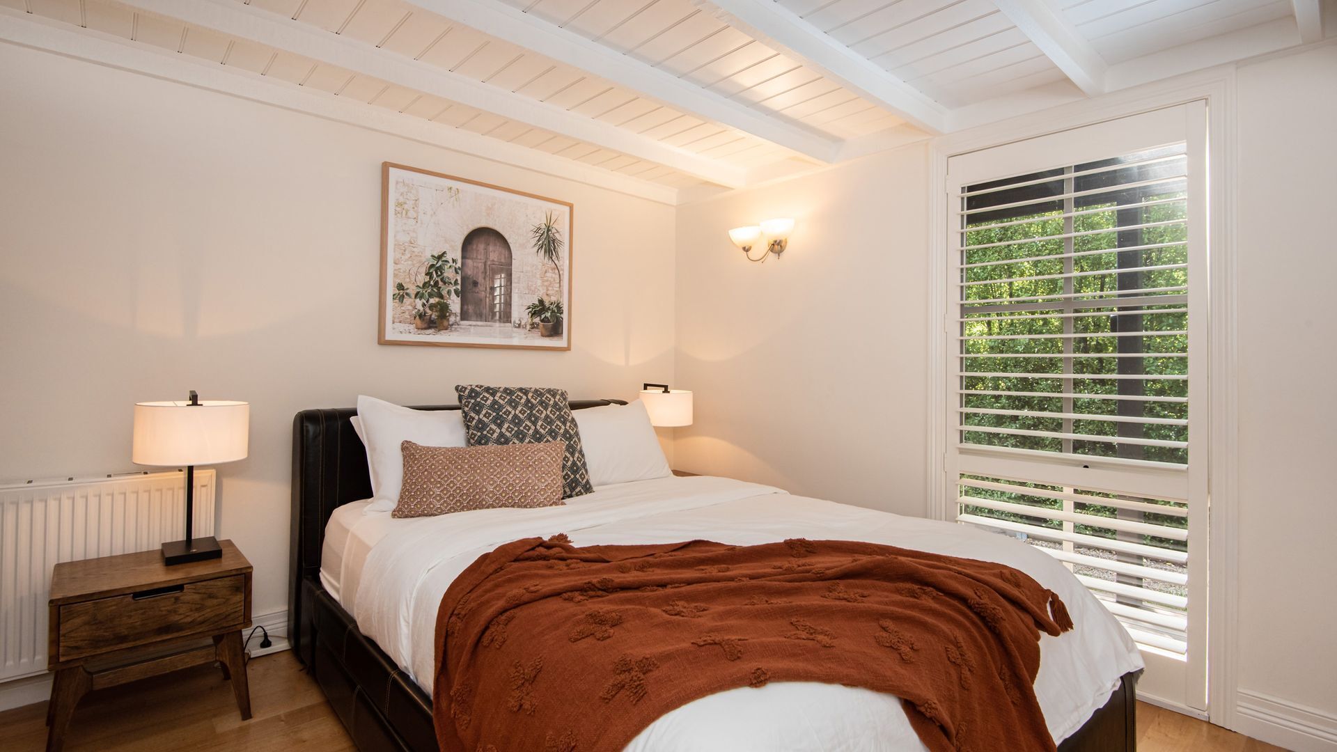 Een slaapkamer met een bed, nachtlampen, een nachtkastje, een raam met jaloezieën en een aan de muur hangend schilderij.