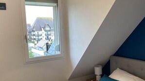 Ein Schlafzimmer mit einem Bett, Kissen, einem Nachttisch, einer Lampe und einem Fenster mit Blick auf Gebäude.
