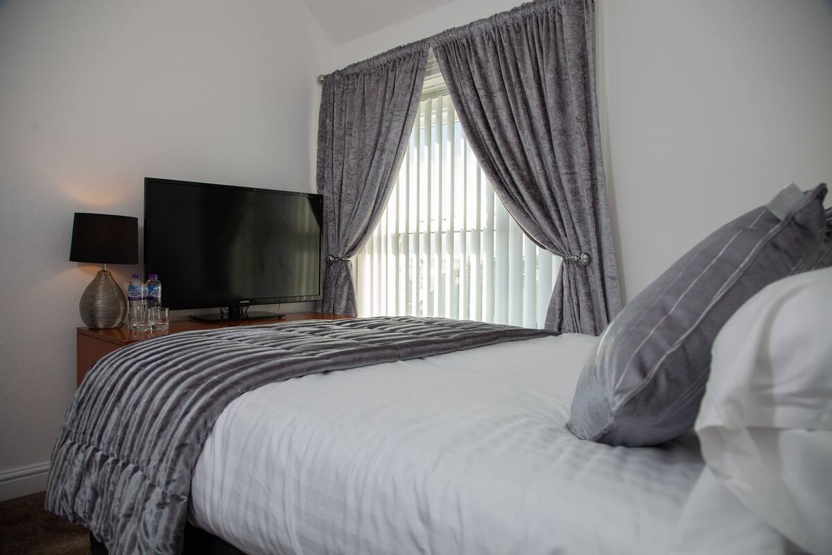 A neatly made bed with a striped blanket, a flat-screen TV on a wooden nightstand, and a window with sheer curtains.