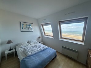 A bedroom with a bed, a nightstand, a lamp, a picture on the wall, and two windows with a beach view. - Apartment with panoramic terrace, pet-friendly, parking (Cabourg)