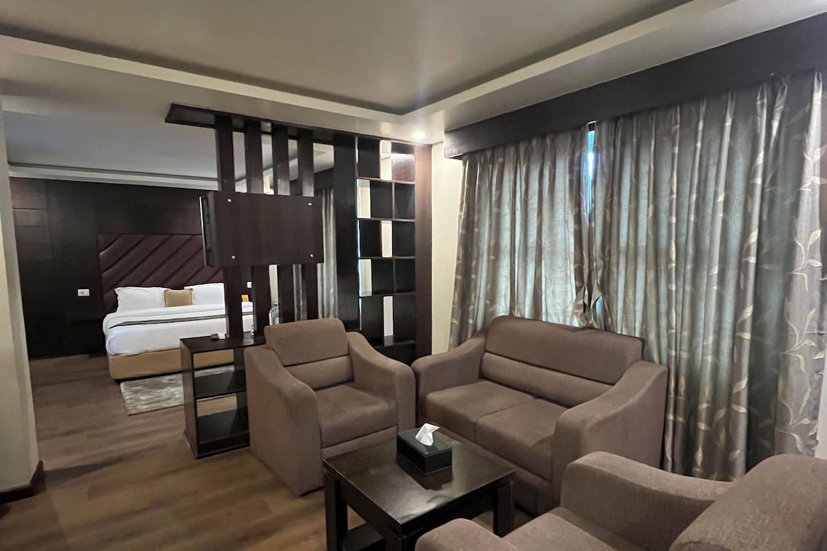 Ein modernes Wohnzimmer mit einer Sofagarnitur, einem Couchtisch und einer TV-Konsole. Im Hintergrund ist ein Bett zu sehen und ein Fenster mit Vorhängen.