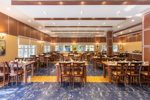 A spacious dining area with multiple tables and chairs, wooden paneling on the walls, and large windows allowing natural light.