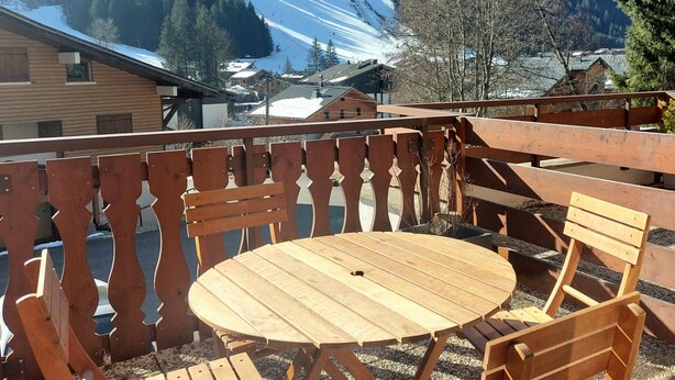 Une terrasse en bois avec une table ronde et des chaises, d’où l’on peut admirer une montagne enneigée et un village.