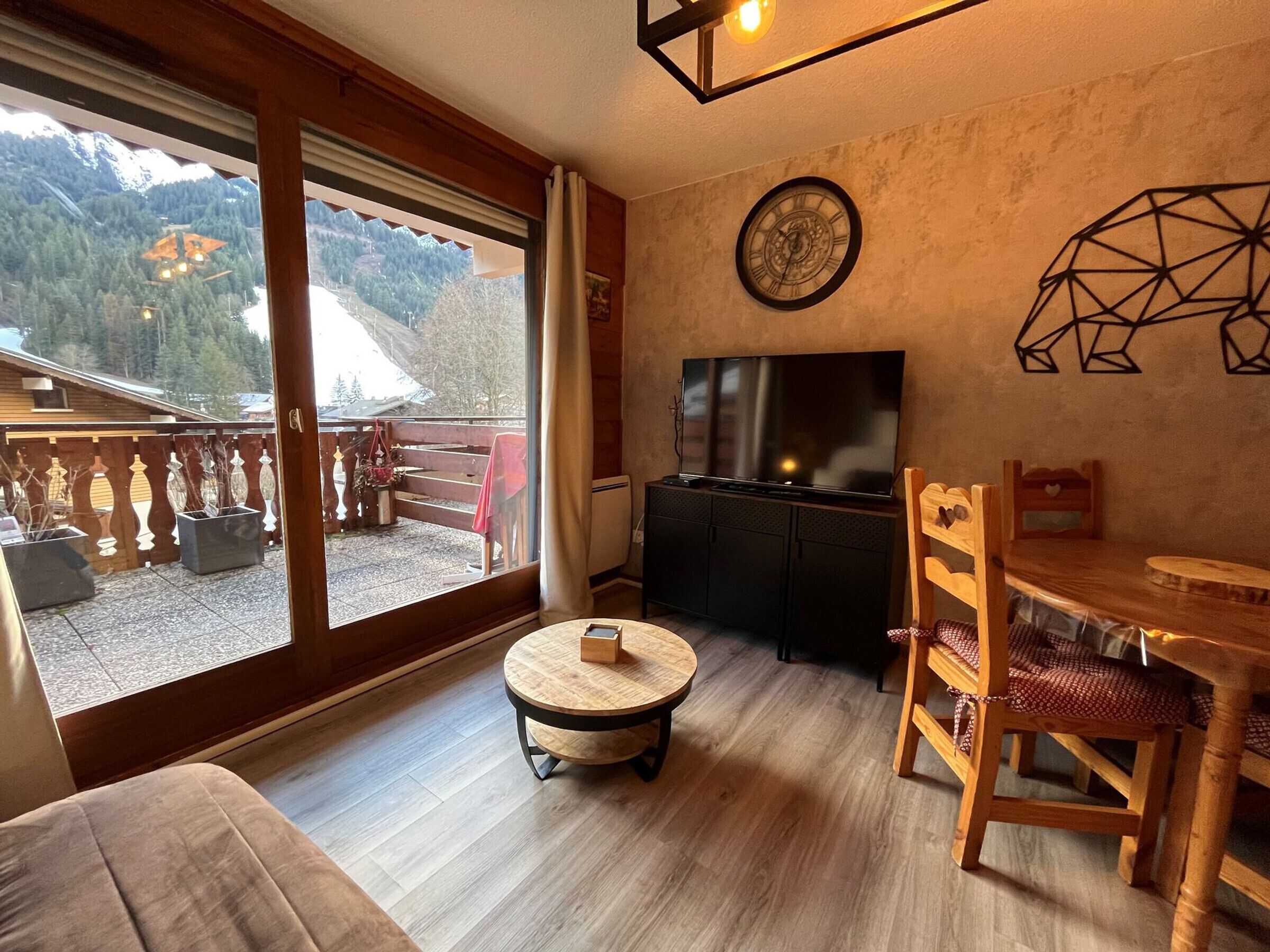A cozy living room with a large sliding glass door leading to a balcony, a round wooden table, a TV on a black cabinet, and a geometric wall decoration.