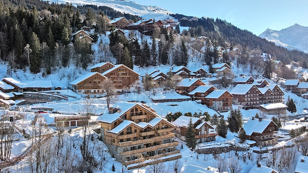 A snow-covered mountain resort with multiple wooden buildings, including a large lodge with balconies, set against a backdrop of snow-capped peaks and dense forest. - Charming Apartment in Méribel - 3 Bedrooms, 6 Pers, Balcony & TV, Services Included (Méribel)