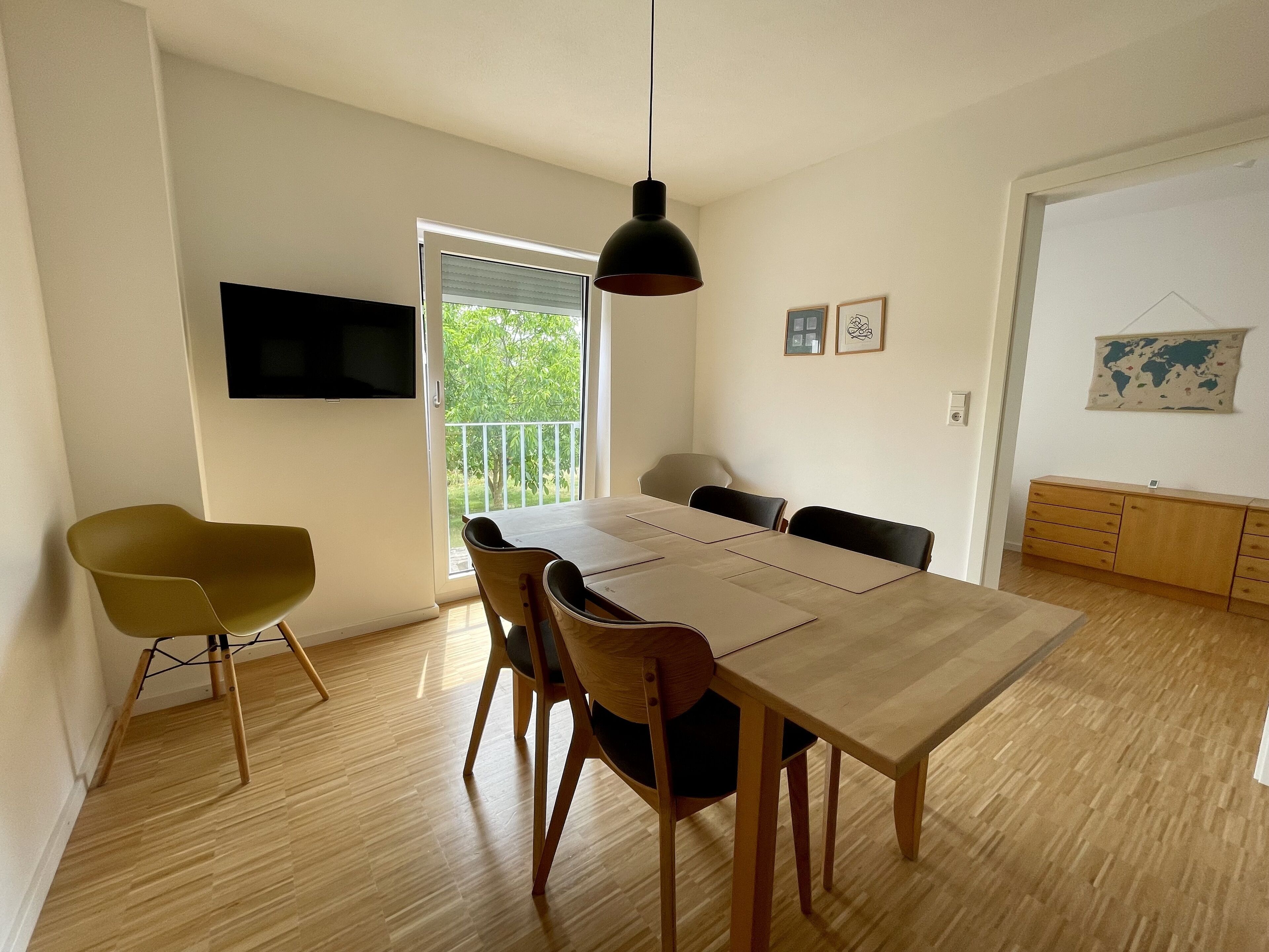 A dining area with a wooden table, chairs, a TV mounted on the wall, and a balcony with a view of greenery.