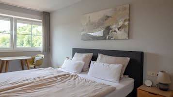 Una cama bien hecha con sábanas blancas, una mesita de noche con lámpara y una silla junto a una ventana con cortinas.