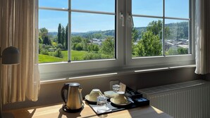 Una habitación con un ventanal grande, un radiador, una mesa con una tetera, tazas y vasos, y una lámpara.