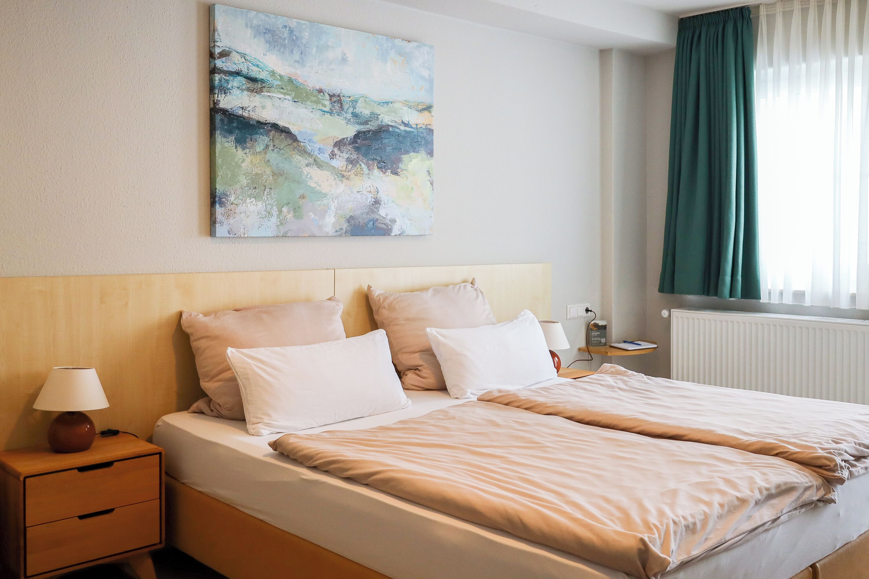 Een hotelkamer met een bed, een nachtkastje, een lamp, een schilderij aan de muur en een raam met gordijnen.