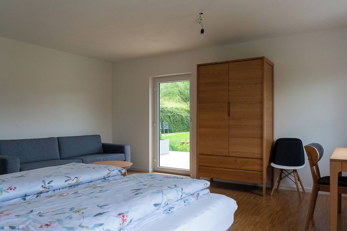 A bedroom with a bed, a grey sofa, a wooden wardrobe, a desk with a chair, and a view of a garden through a door.