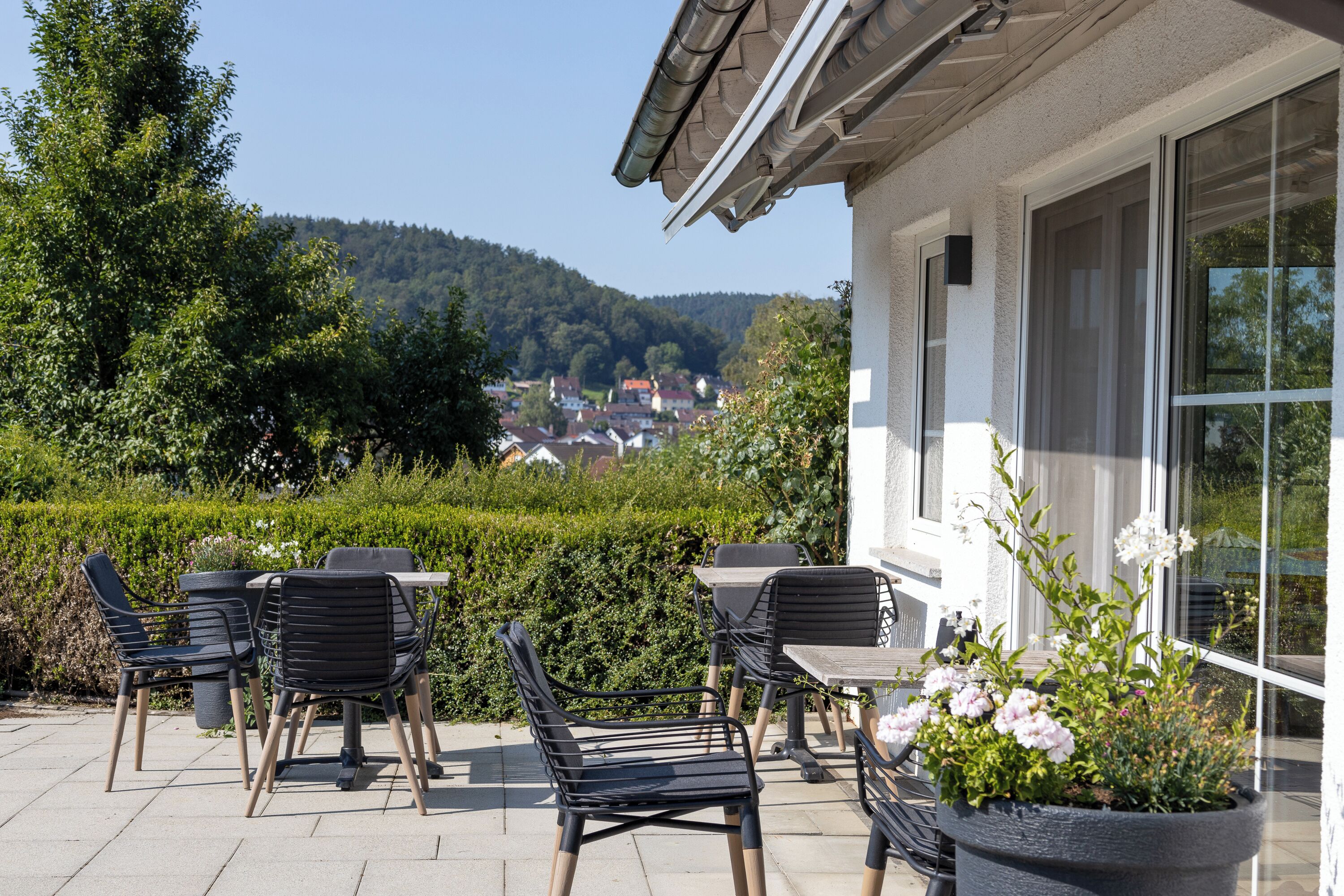 Un patio al aire libre con sillas y mesas, una maceta con flores y vistas a una colina con casas.