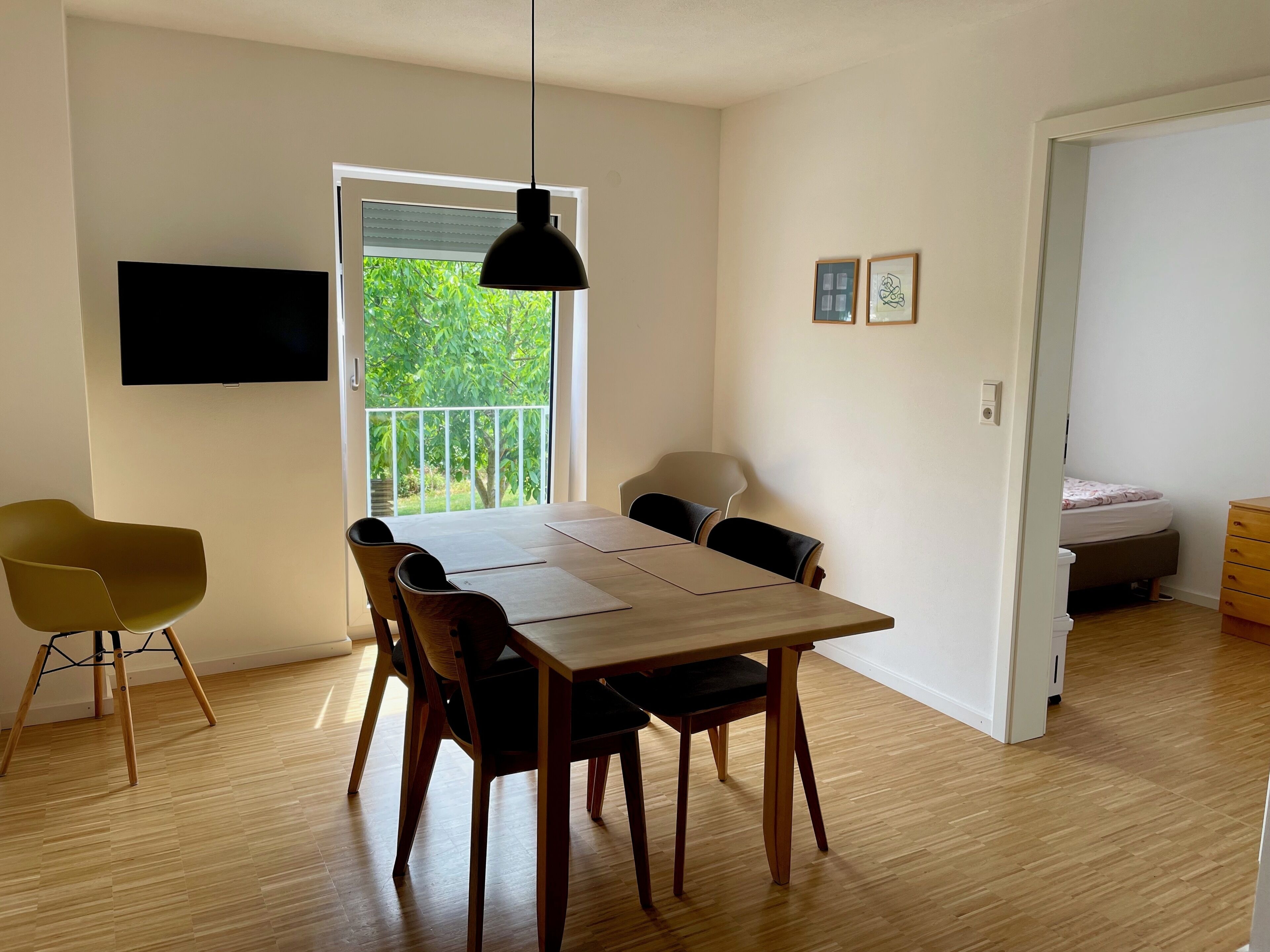 Un comedor con mesa y sillas, un televisor montado en la pared y un balcón con vistas a zonas verdes.