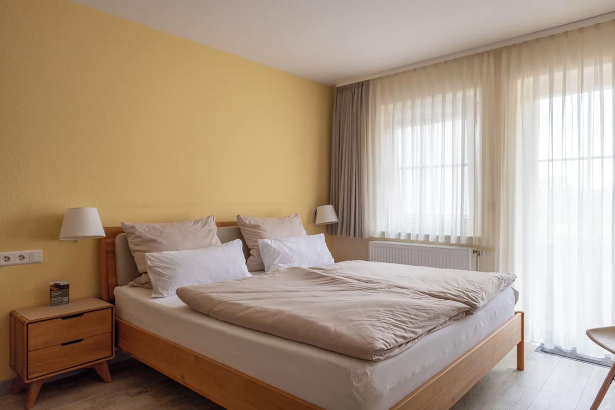 A neatly made bed with a wooden frame, two bedside tables with lamps, a window with sheer curtains, and a radiator.
