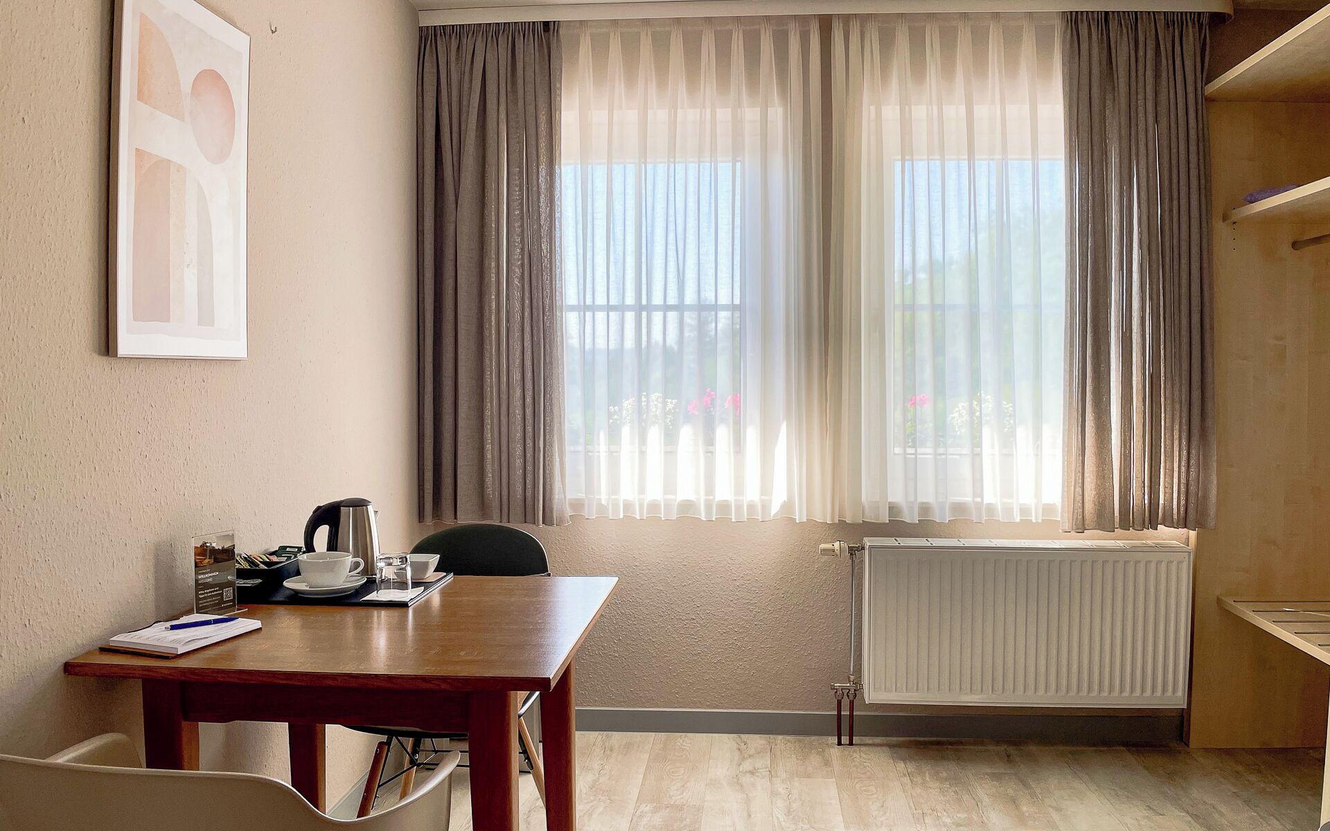 A room with a wooden table, a kettle, a cup, and a chair. There is a radiator on the wall and a window with curtains.