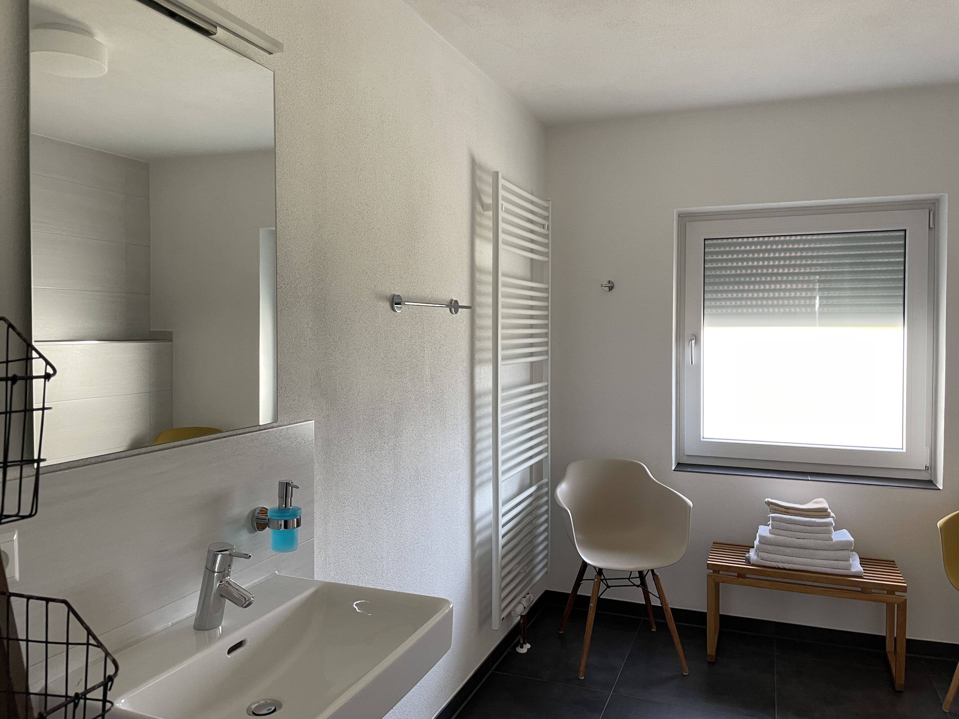 A modern bathroom with a large mirror, a sink, a chair, and a window with blinds.