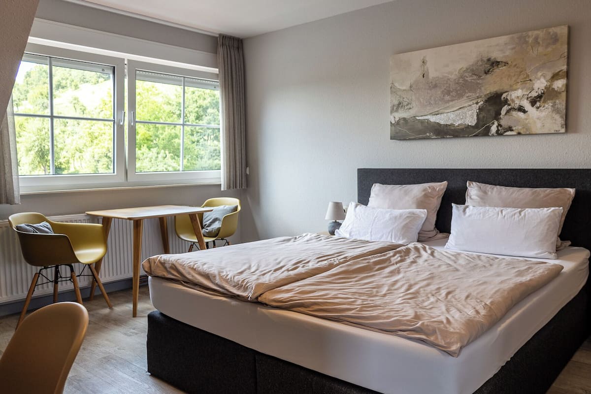 A modern bedroom with a large bed, a small table, two chairs, and a nightstand with a lamp.