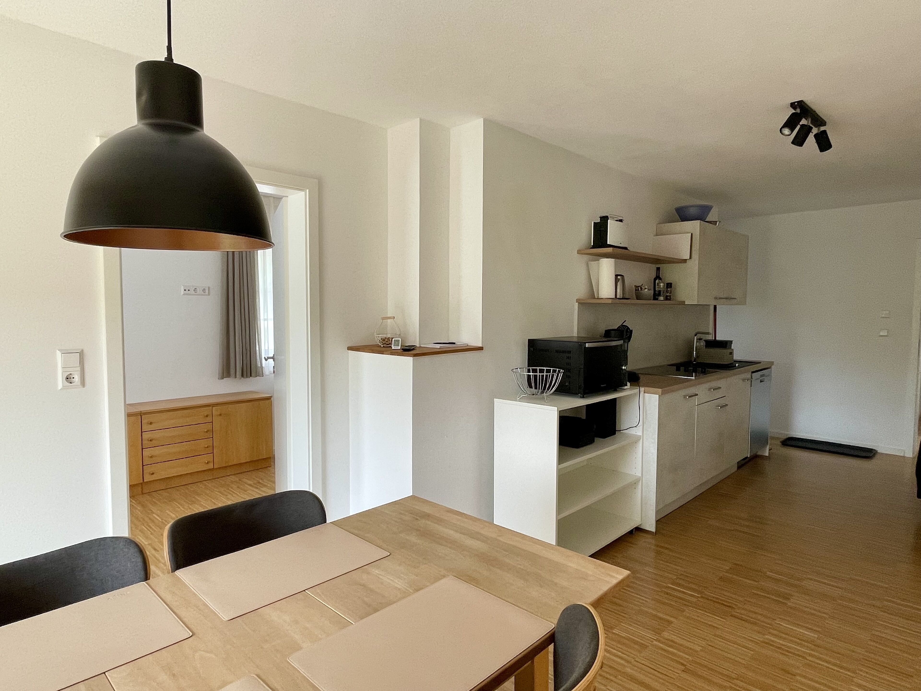 Une cuisine moderne avec des meubles blancs, une table à manger en bois et un luminaire suspendu noir.