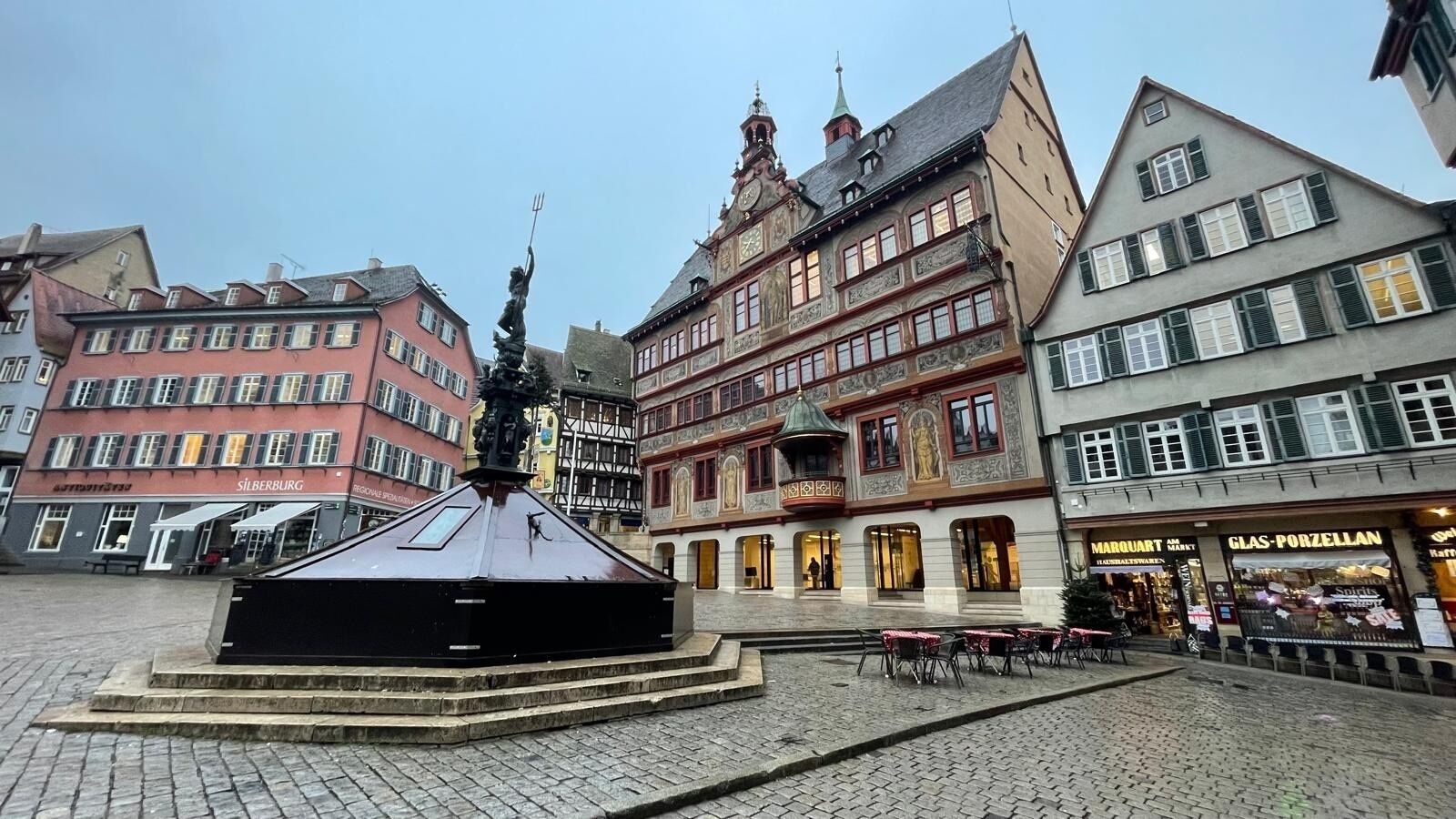 Een historisch plein met een centrale fontein, omringd door traditionele Europese gebouwen met winkels en cafés.