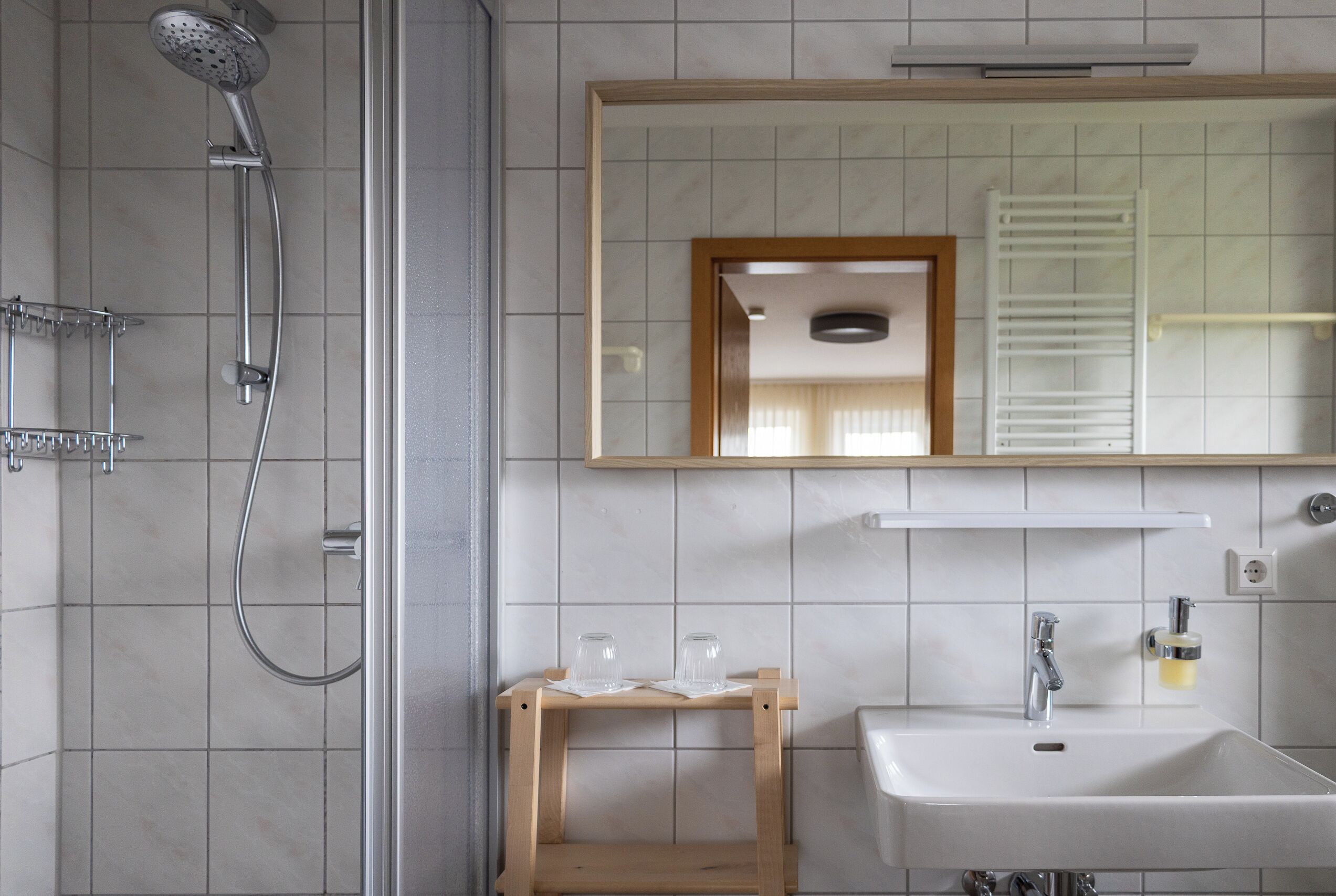 Een badkamer met een douche, wastafel, spiegel en een klein houten krukje.