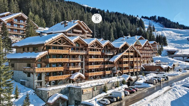 A multi-story wooden lodge with balconies, surrounded by snow-covered trees and a ski slope.