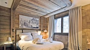 A bedroom with a wooden ceiling, a large bed with white linens, a nightstand with a lamp, and a window with curtains.