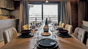 A dining area with a long table set for a meal, wooden ceiling, and a view of snowy mountains through large windows.