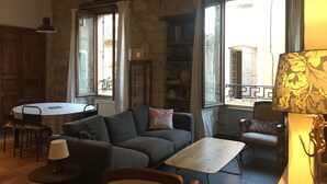 A cozy living room with a sofa, a small table, and a lamp. There are windows with curtains, a bookshelf, and a dining area with chairs. - In a historic 12th-century building (Montfrin)