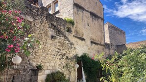 A stone building with a rustic charm, a small outdoor seating area with metal chairs and a table, surrounded by potted plants and flowering shrubs. - In a historic 12th-century building (Montfrin)