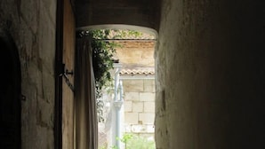 A narrow corridor with a small bench, a door with a lock, and a view of a courtyard with potted plants and a tiled roof. - In a historic 12th-century building (Montfrin)