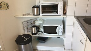 Une cuisine compacte avec des appareils électroménagers blancs, notamment un grille-pain, une machine à café et un four à micro-ondes. On y trouve une poubelle et une étagère avec une horloge.