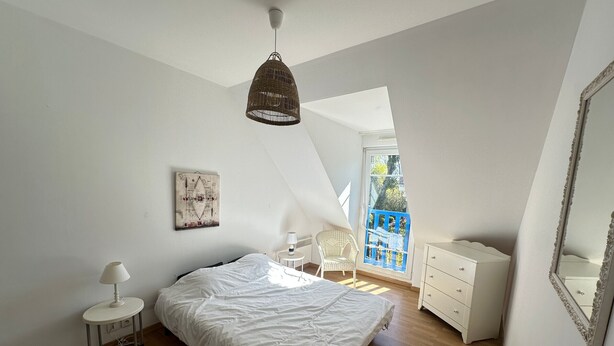 Une chambre à coucher comprenant un lit, une commode blanche, une chaise, une lampe, un luminaire suspendu, un tableau accroché au mur et une fenêtre avec un garde-corps bleu.