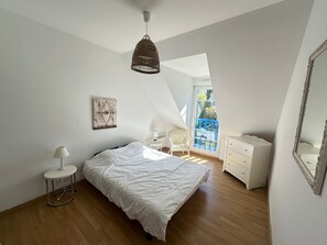 A bedroom with a bed, a white dresser, a chair, a lamp, a hanging light fixture, a wall-mounted picture, and a window with a blue railing. - Spacious Villa Near Beach and Town Center with Large Private Garden (Villers-sur-Mer)