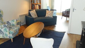 A living room with a blue sofa, a patterned armchair, a wooden coffee table, and a white chair. There is a bookshelf with books and a lamp. - Cabourg: Modern 4/5-room apartment, beach access, 8 ppl, garden and terrace, parking, WIFI (Cabourg)