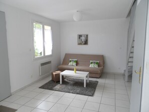 Una habitación pequeña y bien iluminada con un sofá color beige, una mesita de café blanca y una alfombra pequeña. Se observa una ventana con cortinas, un radiador y una puerta que da a una escalera.