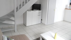 A modern, minimalistic living space with a staircase, a flat-screen TV on a cabinet, and a small table with a vase. - Charming 3-Room Cottage with Private Garden, Near Marina, Sleeps 6, Pet-Friendly, Pool Access (Dives-sur-Mer)
