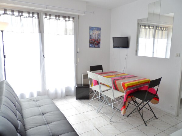 Ein Wohnzimmer mit grauem Sofa, einem Esstisch mit bunten Tischdecken, einem an der Wand montierten Fernseher und einem Fenster mit Vorhängen.