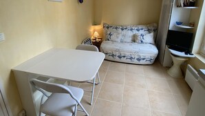 Habitación pequeña con cama, una mesita y una silla. Se observa una estantería con objetos decorativos y un televisor.