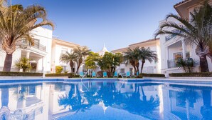 Une piscine à l’eau d’un bleu cristallin, entourée de palmiers et de bâtiments blancs dotés de balcons.