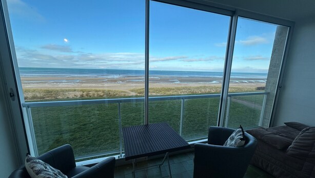 A room with a large window offering a view of a beach and grassy area. There is a small table and two chairs. - Stunning Sea View Apartment in Varaville – Beachfront Loggia, Sleeps 4 (Le Home Varaville)