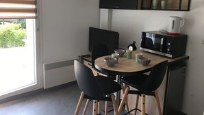 A compact kitchen with a microwave, kettle, and a coffee maker on a shelf. There are two black chairs around a small wooden table.