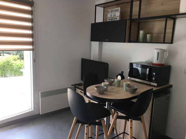 A modern kitchen with a microwave, kettle, and a coffee maker on the counter. There are two black chairs and a small table with bowls and cups. A sign on the wall reads 'BIENVENUE A LA MER.'