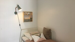 A small, neatly arranged bedroom with a single bed, a bedside lamp, a wall-mounted storage unit, and a power outlet on the wall.