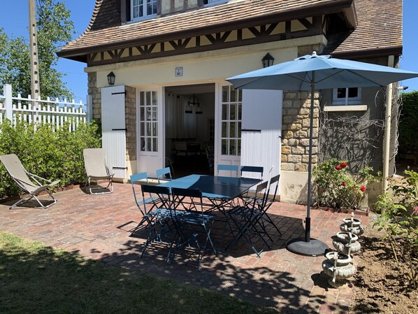 A house with a patio featuring a table and chairs, a white fence, and a garden with plants and a bird bath.